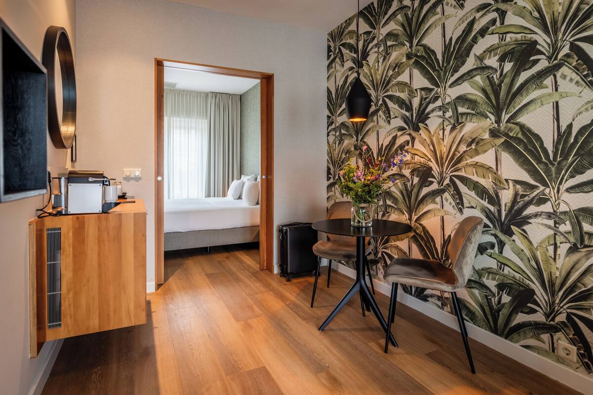 Een compacte hotelkamer met een houten vloer, een bed met witte lakens, een tafeltje met een bloemenvaas en een muur met een tropisch bladmotief.