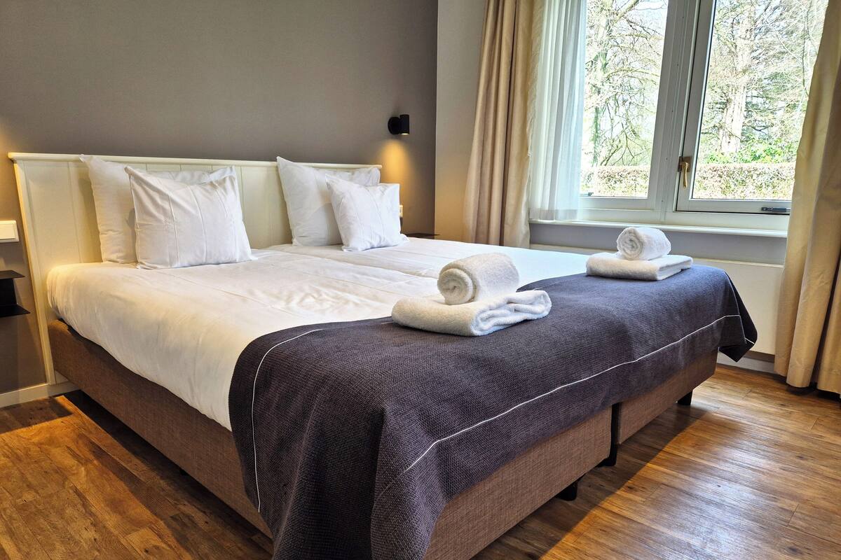 Een tweepersoonsbed met witte lakens en een donkerblauwe deken, een bedlampje en een raam met gordijnen.