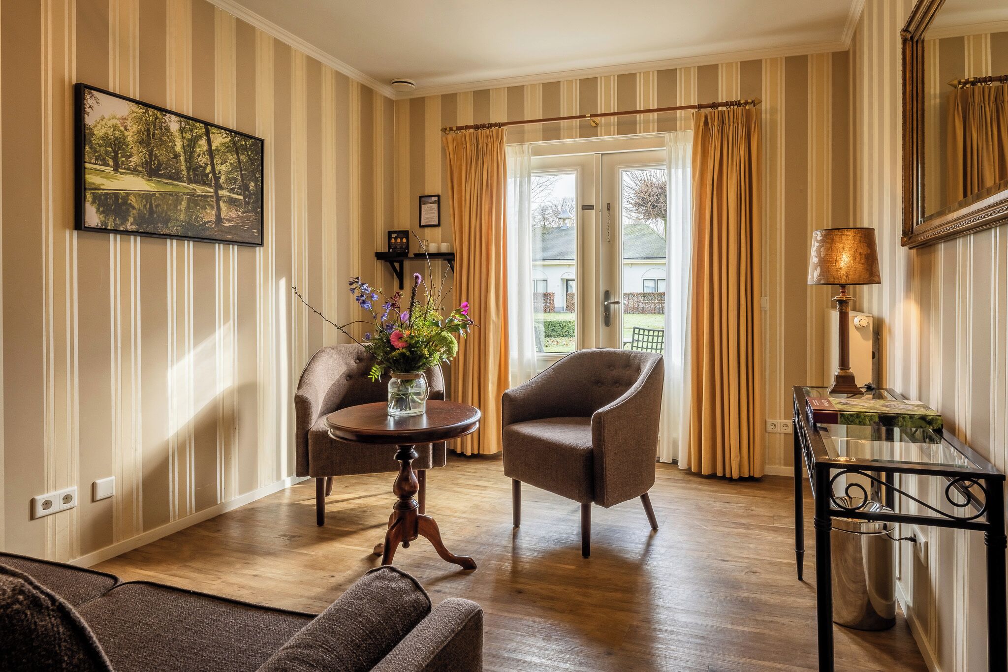 A room with striped wallpaper, a wooden floor, a sofa, a chair, a table with a vase of flowers, a lamp, and a door leading outside.