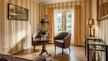 A room with striped wallpaper, a wooden floor, a sofa, a chair, a table with a vase of flowers, a lamp, and a door leading outside.