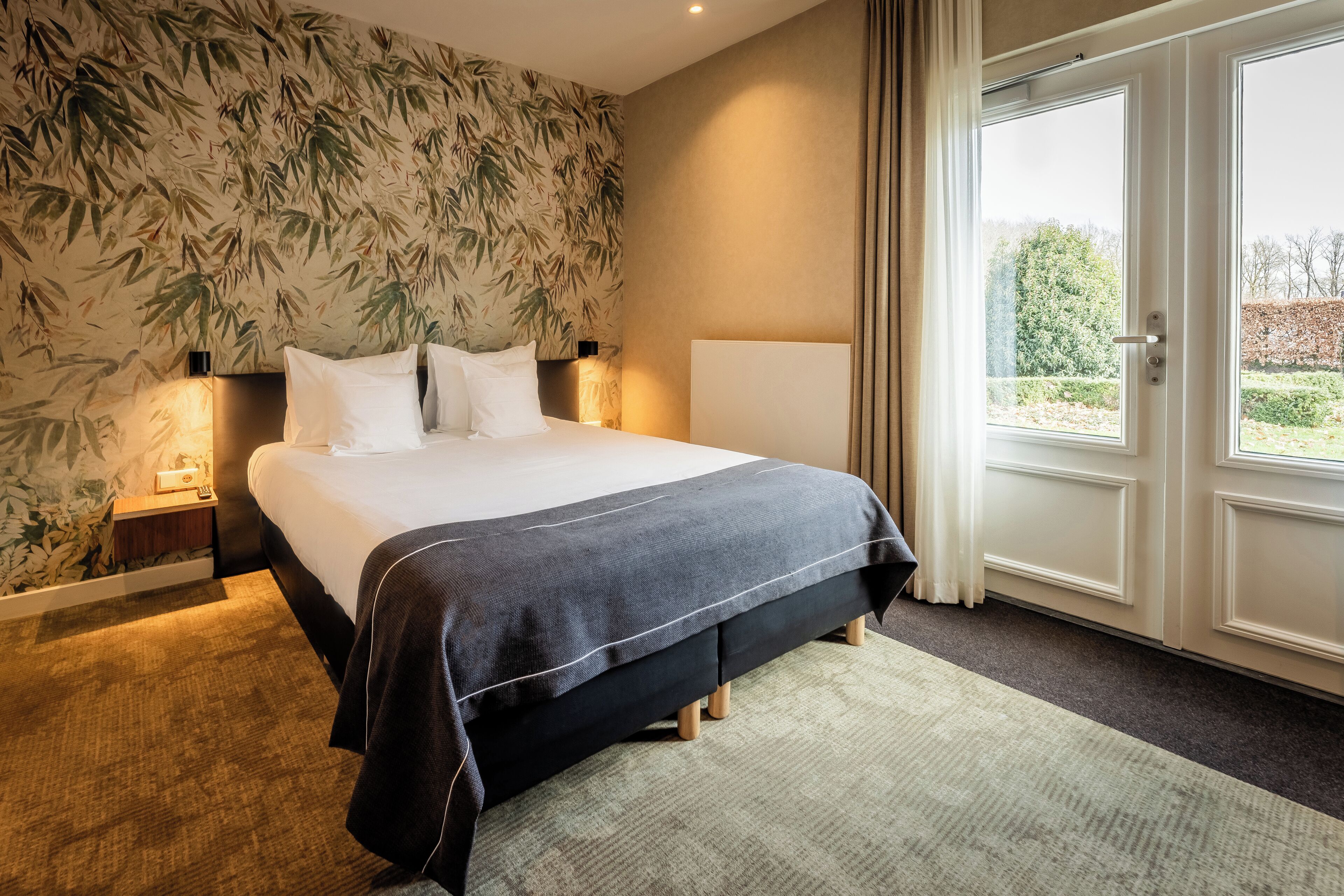 Hotelkamer met een bed, nachtkastje, lamp, raam met gordijnen en een wand met bamboeblad behang.