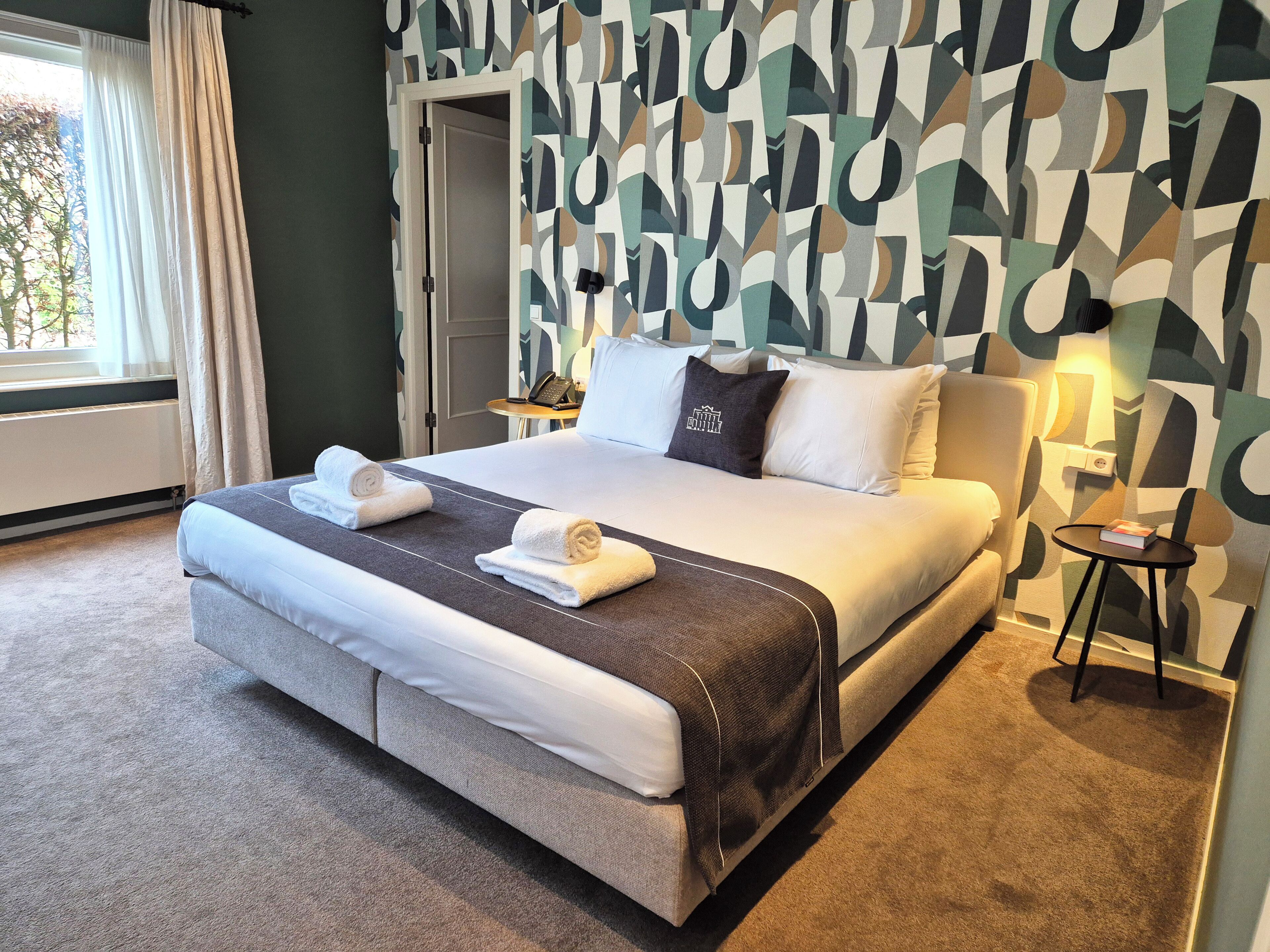 A neatly made bed with towels, a bedside table with a lamp, and a patterned wall.