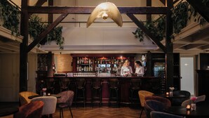 Zona de bar con vigas de madera, luces colgantes tipo lámpara y taburetes. Hay mostradores con un cantinero y estanterías llenas de botellas.