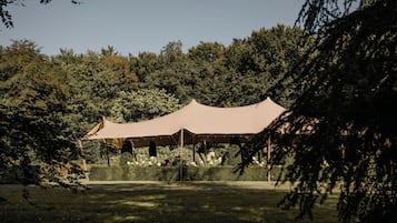 A large, open tent structure in a park-like setting with dense trees in the background.