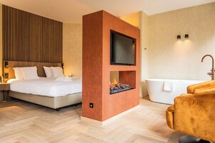 Una habitación de hotel moderna con una cama grande, una chimenea integrada, un televisor, una bañera y una zona de estar.
