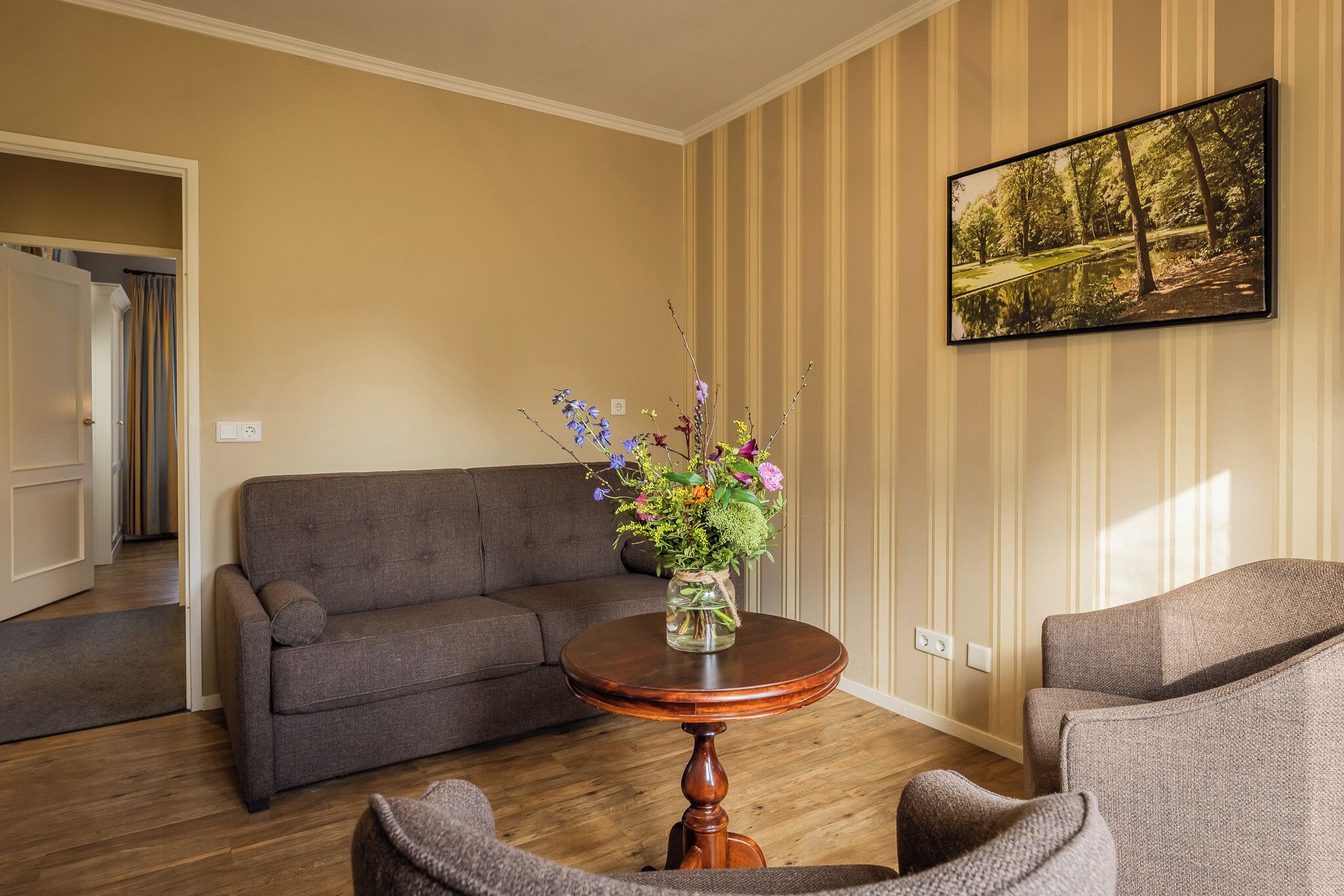 Una sala de estar con un sofá, sillones, una mesa redonda de madera con un jarrón de flores y un cuadro en la pared.
