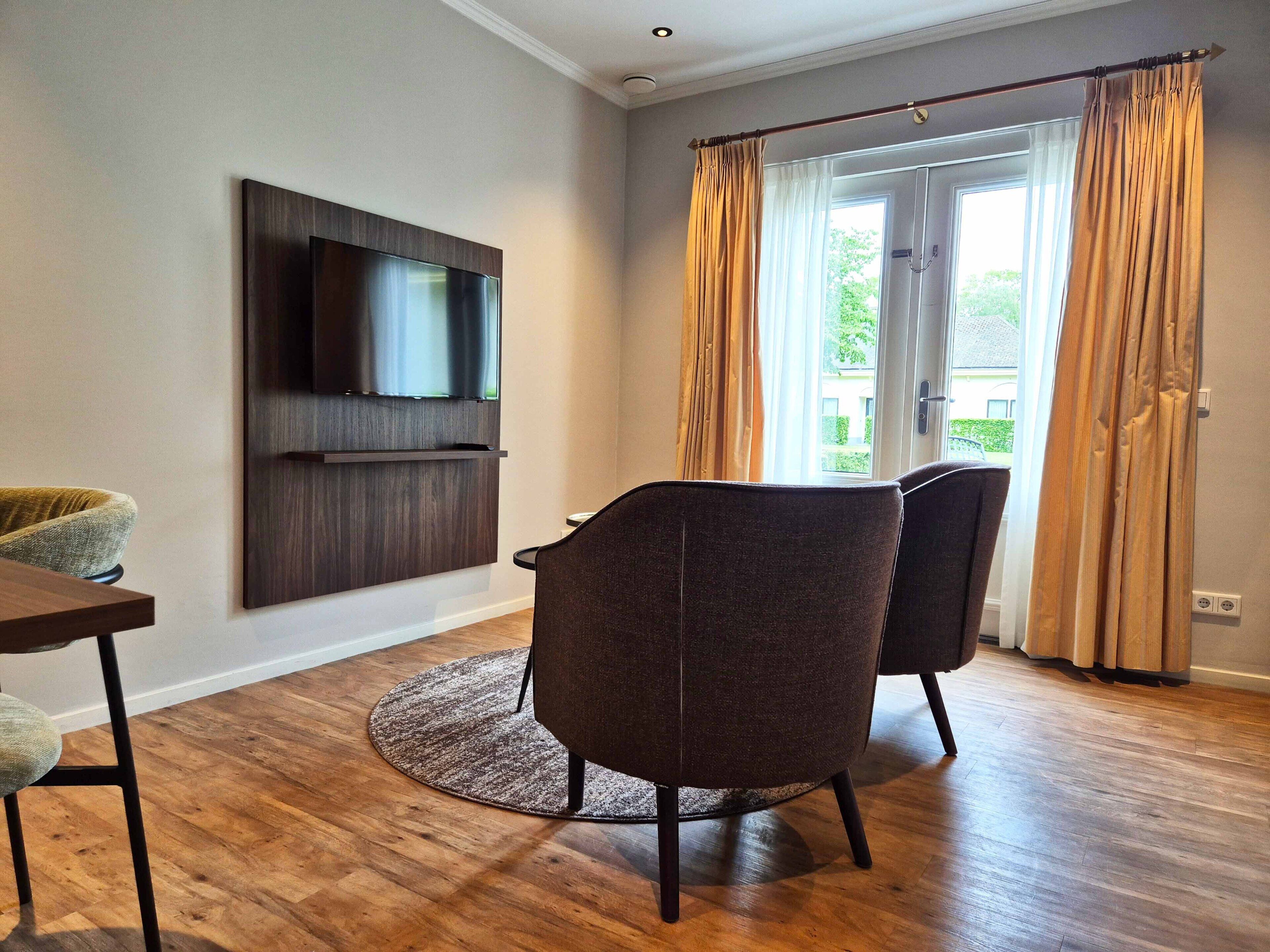 A modern living room with a flat-screen TV mounted on the wall, two dark chairs, a round rug, and a wooden floor.