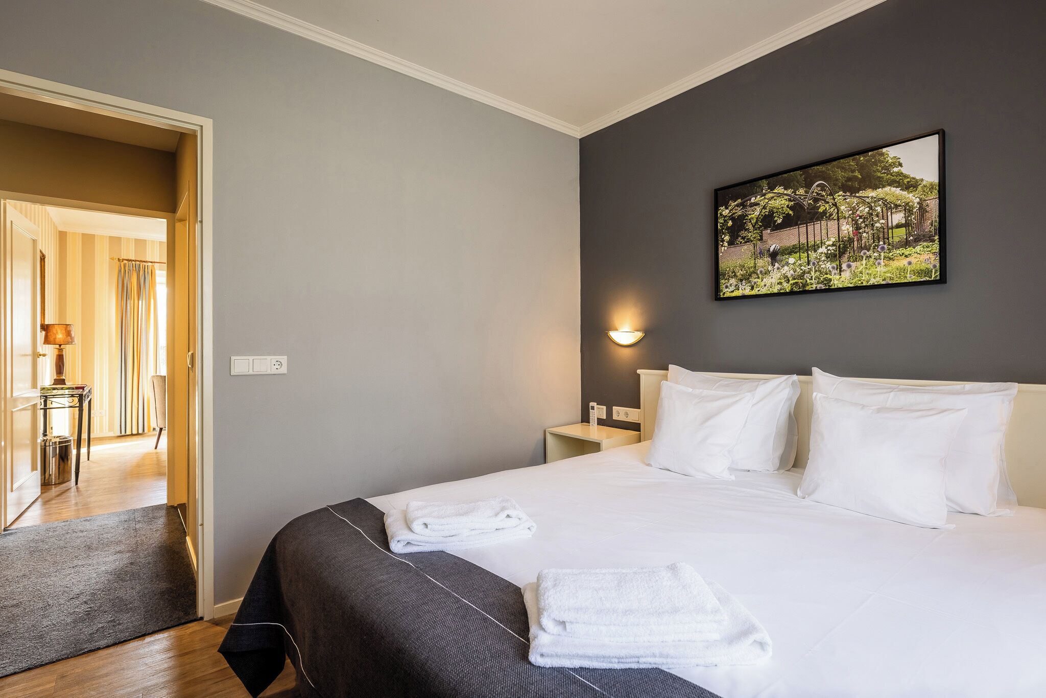 A hotel room with a bed, a nightstand, a wall-mounted picture, and a door leading to another room.