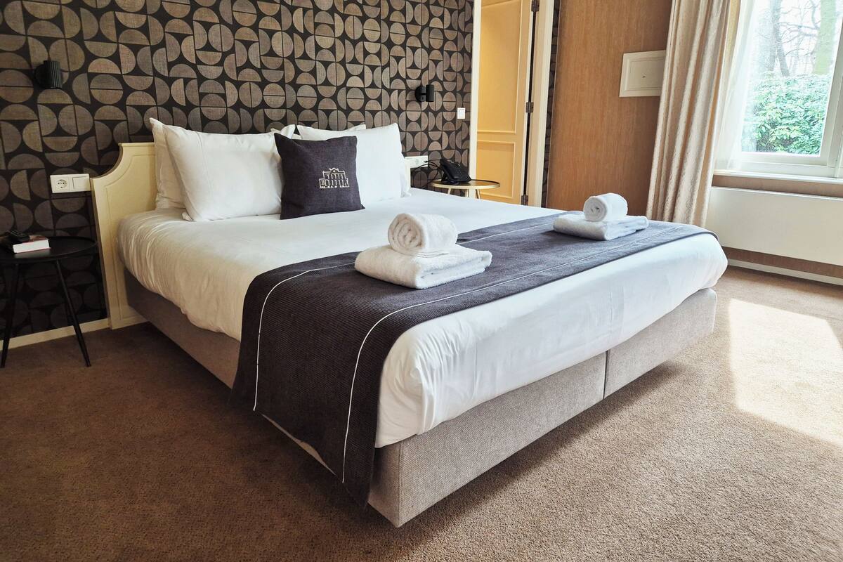 Een opgemaakt bed met witte lakens en een donkere plaid, een klein nachtkastje met een boek en een telefoon, en een behang met een patroon.