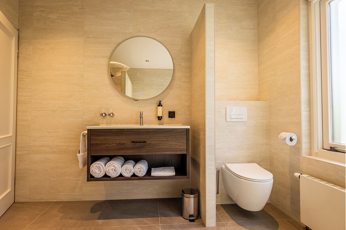 Un bany modern amb un lavabo de fusta, un mirall rodó, un vàter i una finestra.