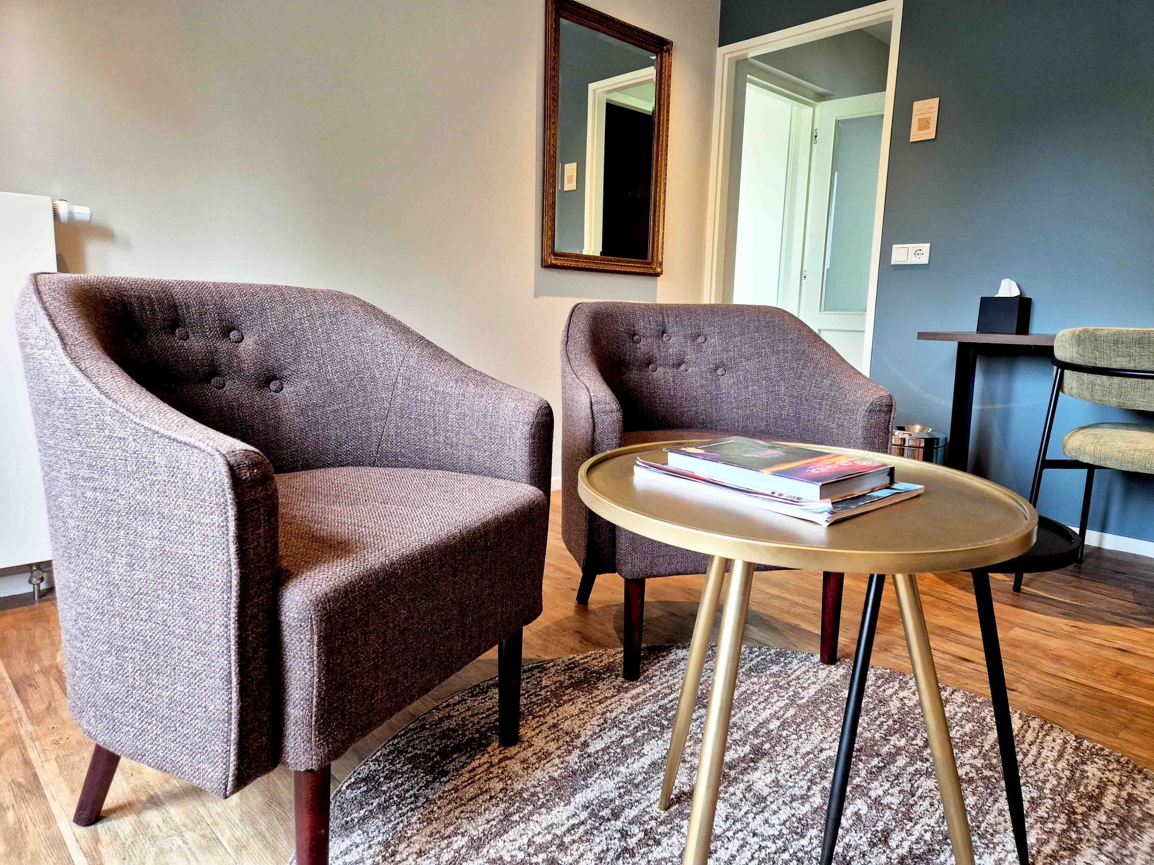 Een kamer met twee fauteuils, een klein rond tafeltje met boeken, een spiegel en een deur.
