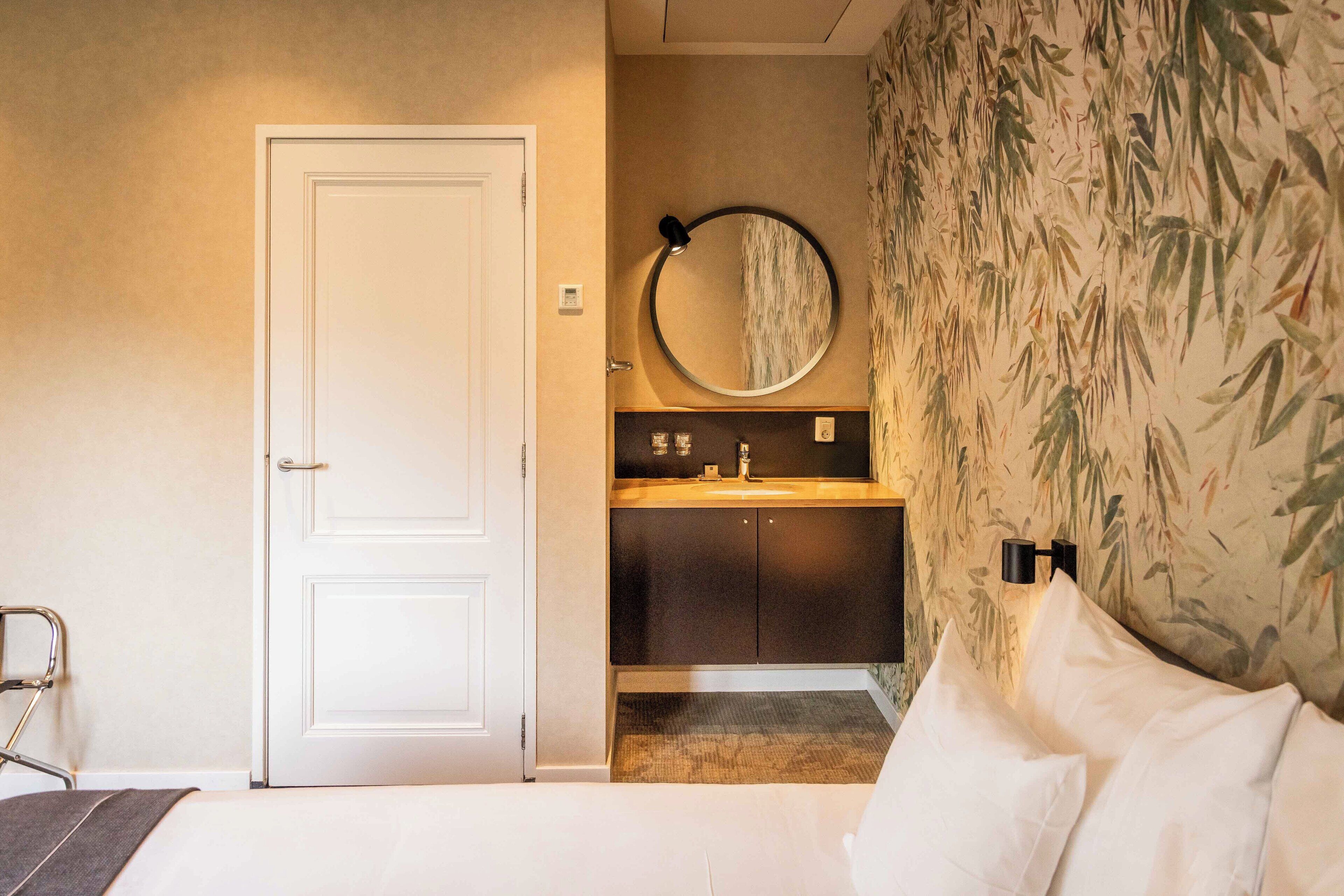 Habitación de hotel con una cama, una puerta blanca, un baño con lavamanos y espejo, y una pared con papel tapiz de bambú.