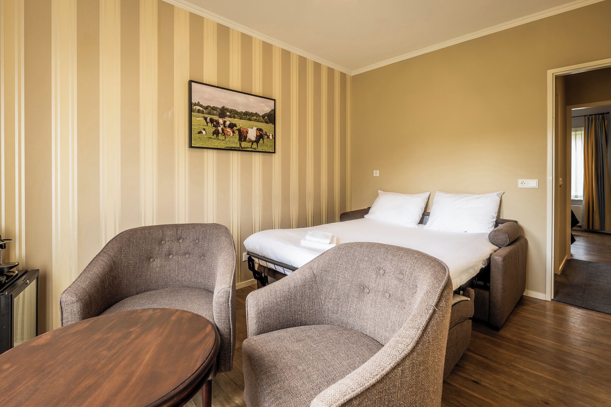 A hotel room with a bed, two armchairs, a wooden table, and a painting of a field with cows.