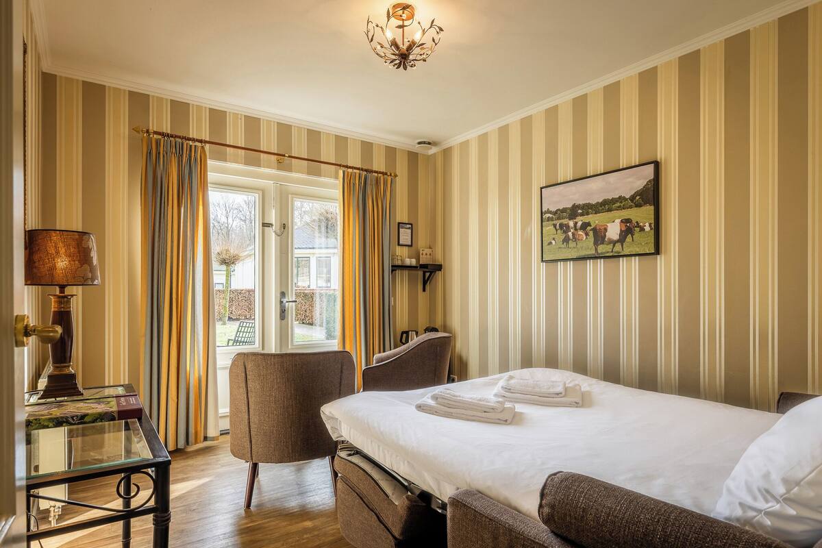 Een hotelkamer met een bed, een stoel, een bureau met een lamp, een schilderij aan de muur en een raam met gordijnen.