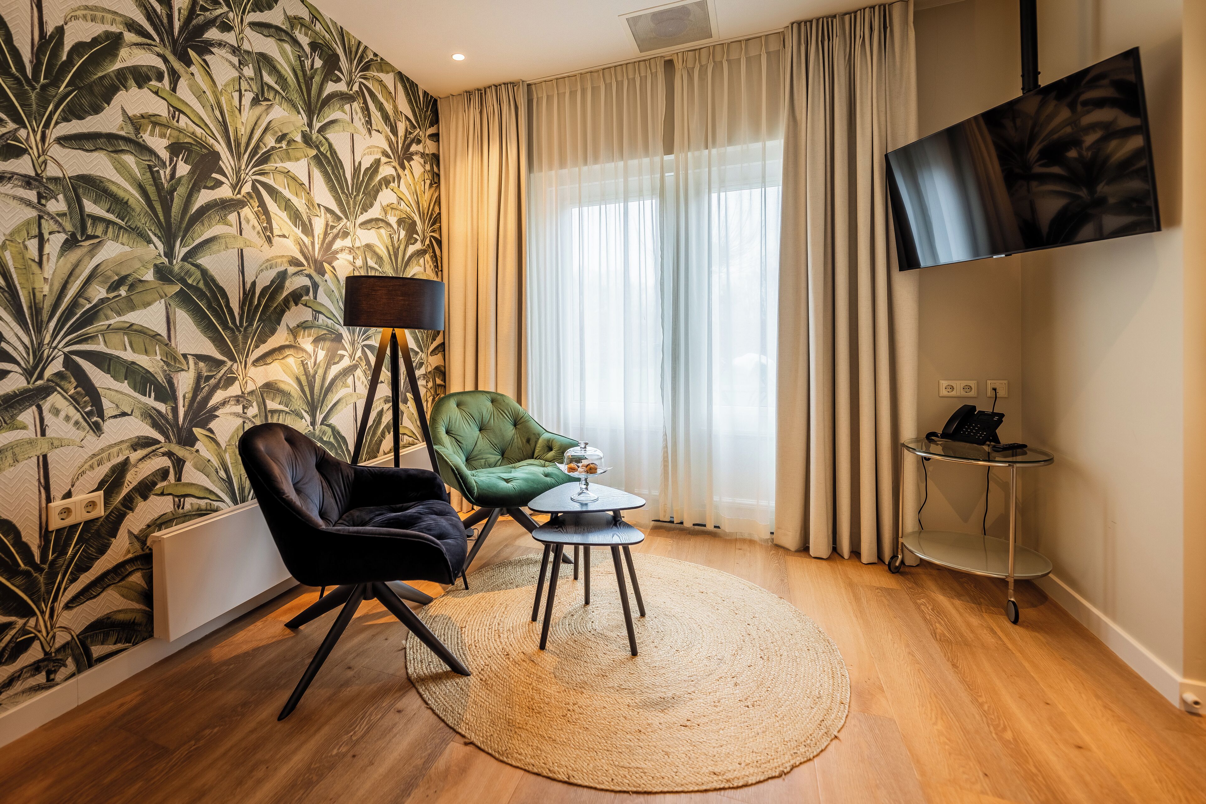 Ein Zimmer mit Holzboden, einem grünen Sessel, einem kleinen runden Tisch, einer Stehlampe, einem an der Wand montierten Fernseher und einem fensterbedeckenden Vorhang.