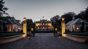 Un edificio grande e histórico con una entrada central flanqueada por dos construcciones más pequeñas, un camino de adoquines y un césped bien cuidado.