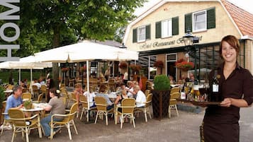 An outdoor restaurant with wicker chairs and white umbrellas, a brick building with a sign reading "Hotel Restaurant," and a waiter serving drinks.