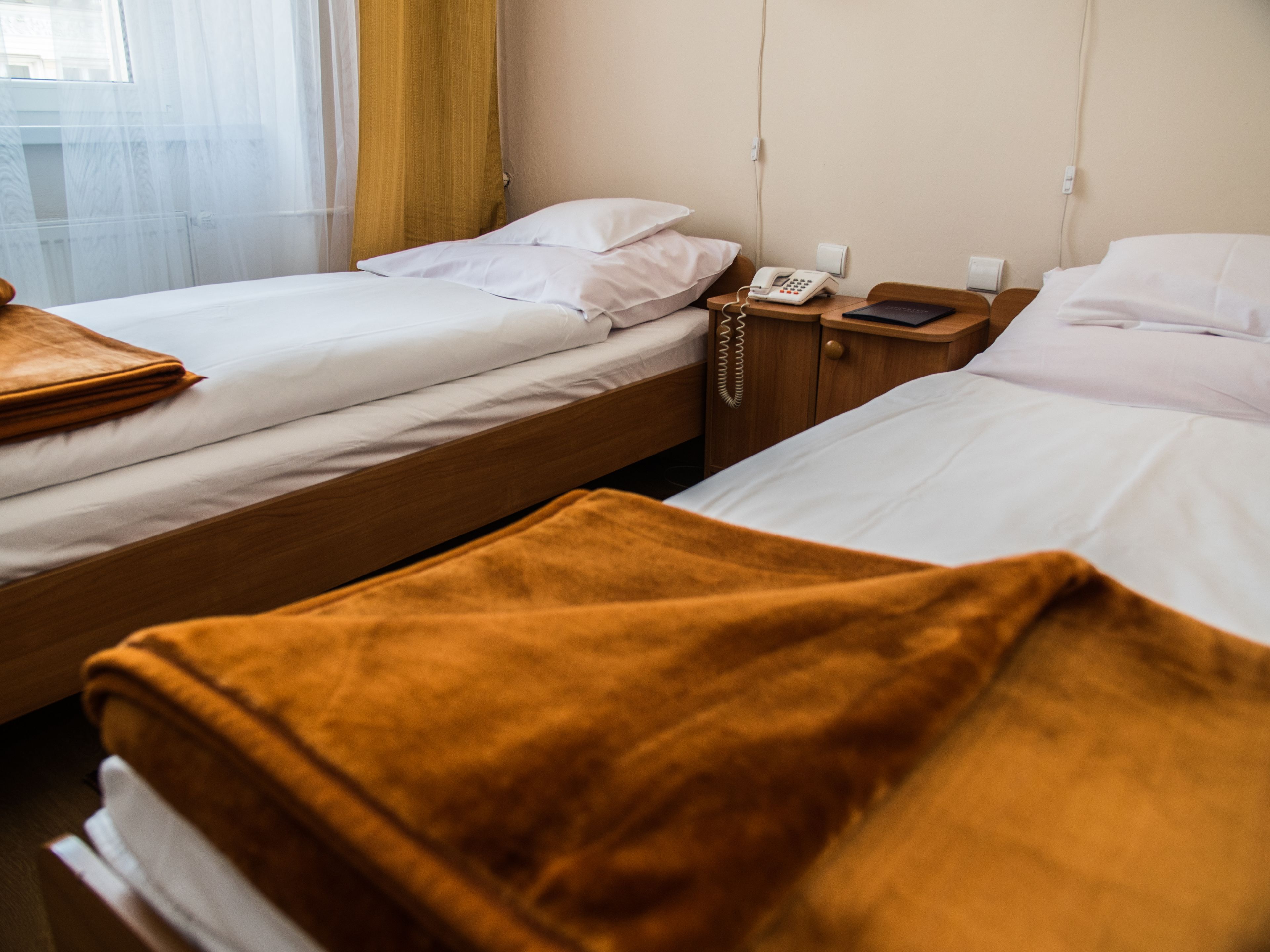 A hotel room with two single beds, a wooden nightstand with a phone and a book, and a window with curtains.