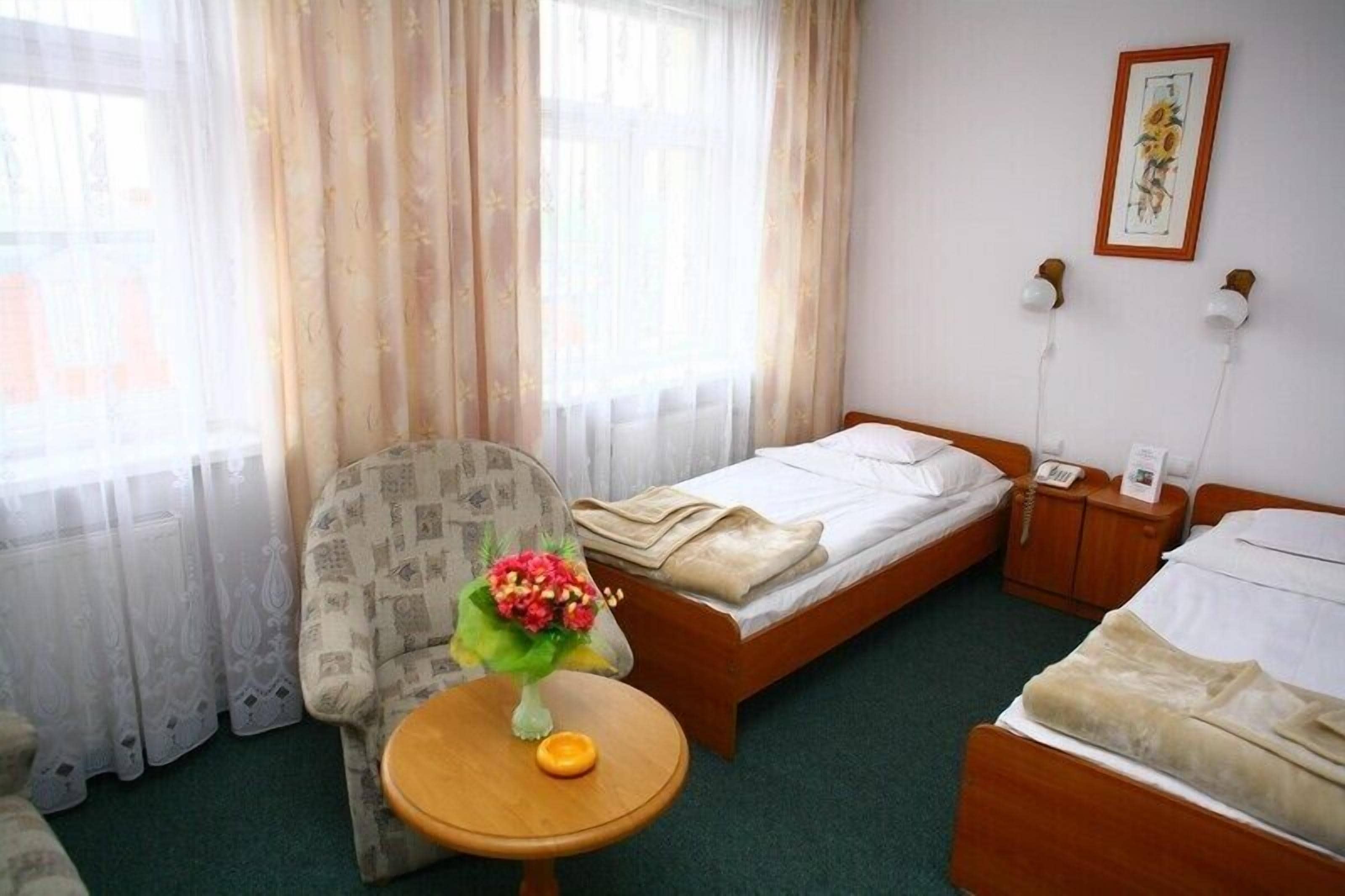 Habitación de hotel con una cama individual, una silla, una mesita con un jarrón de flores y una ventana con cortinas translúcidas.