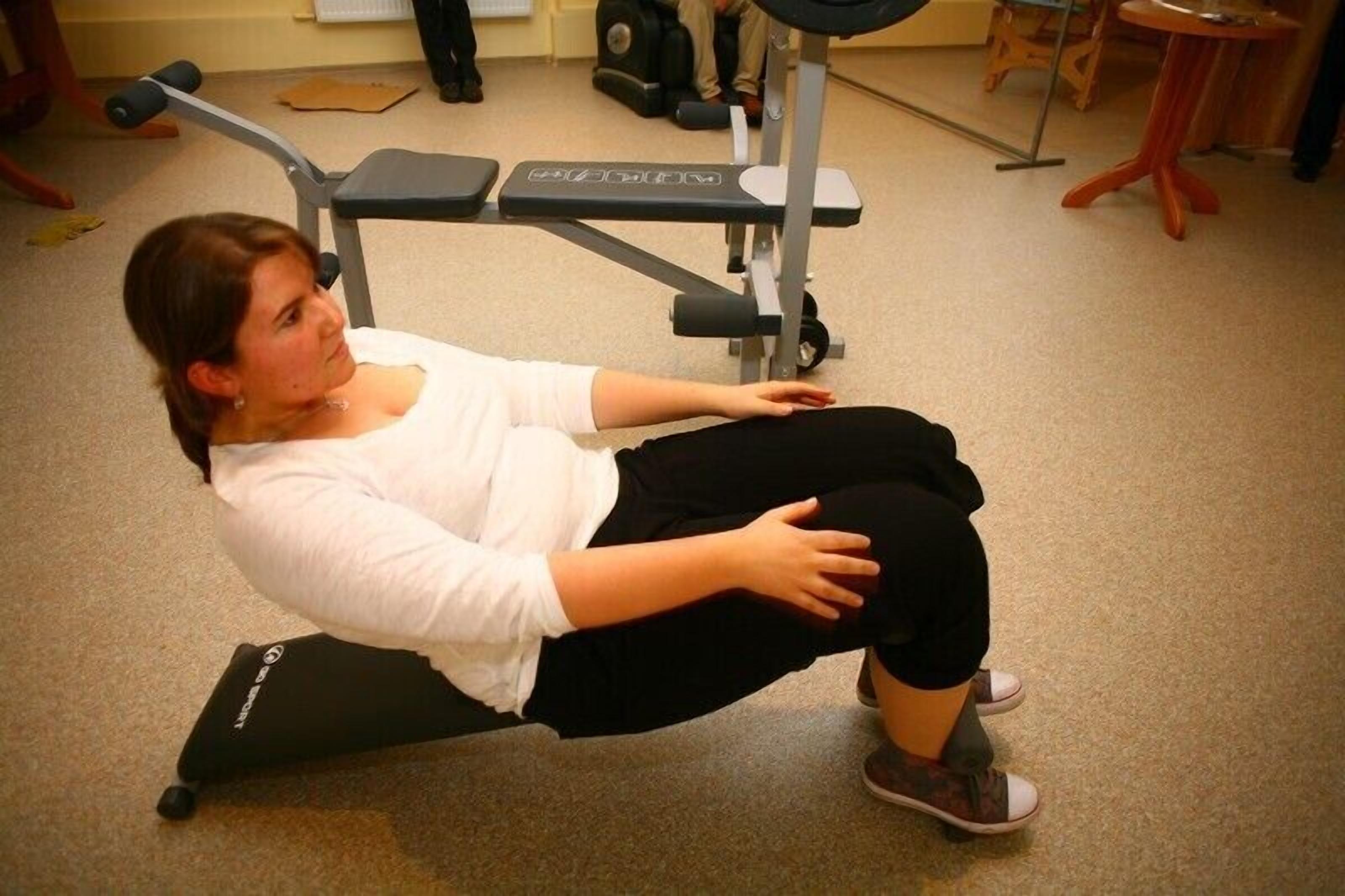 A person seated on an exercise machine in a room with a carpeted floor, a bench, and a chair in the background.