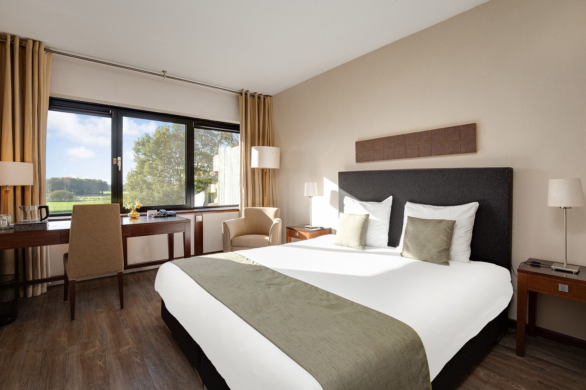 A hotel room with a large bed, a desk, a chair, a nightstand, and a window with curtains.
