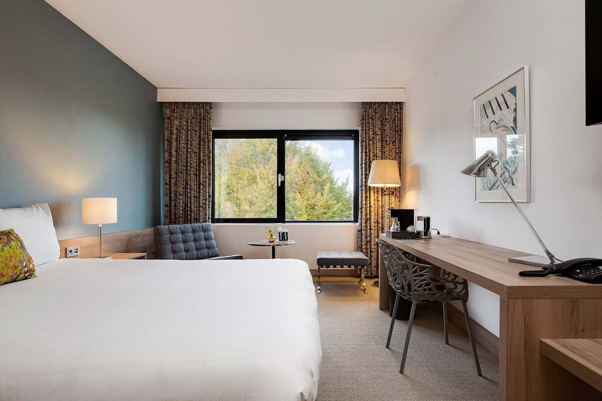 Une chambre d’hôtel avec un lit, un bureau, une chaise, une lampe et une fenêtre avec des rideaux.