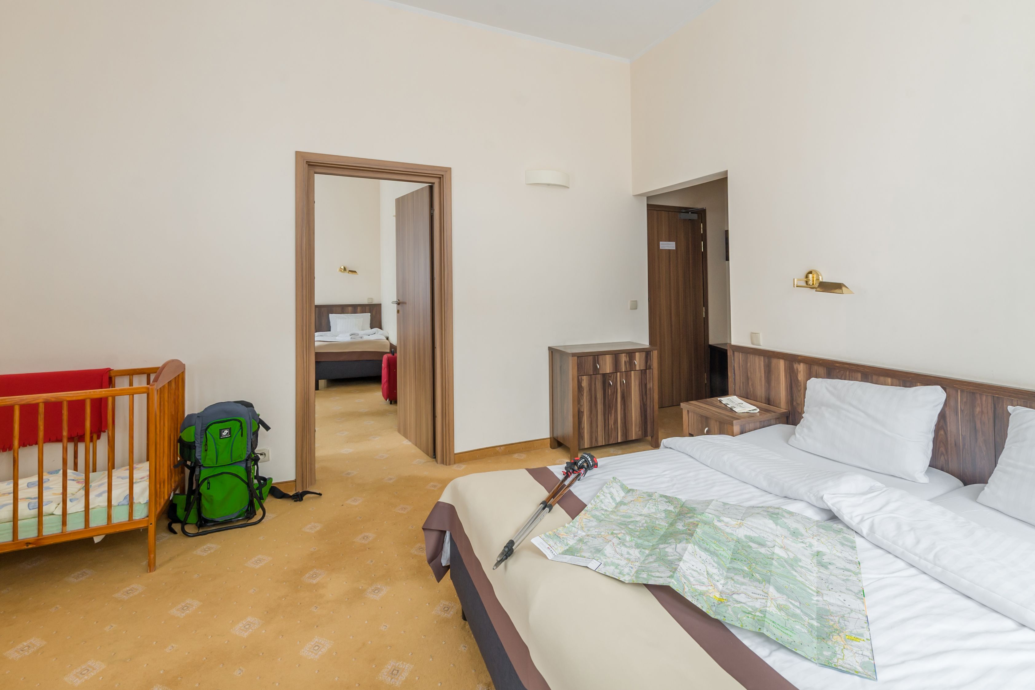 A hotel room with a bed, a crib, a green backpack, a wooden cabinet, and a nightstand.