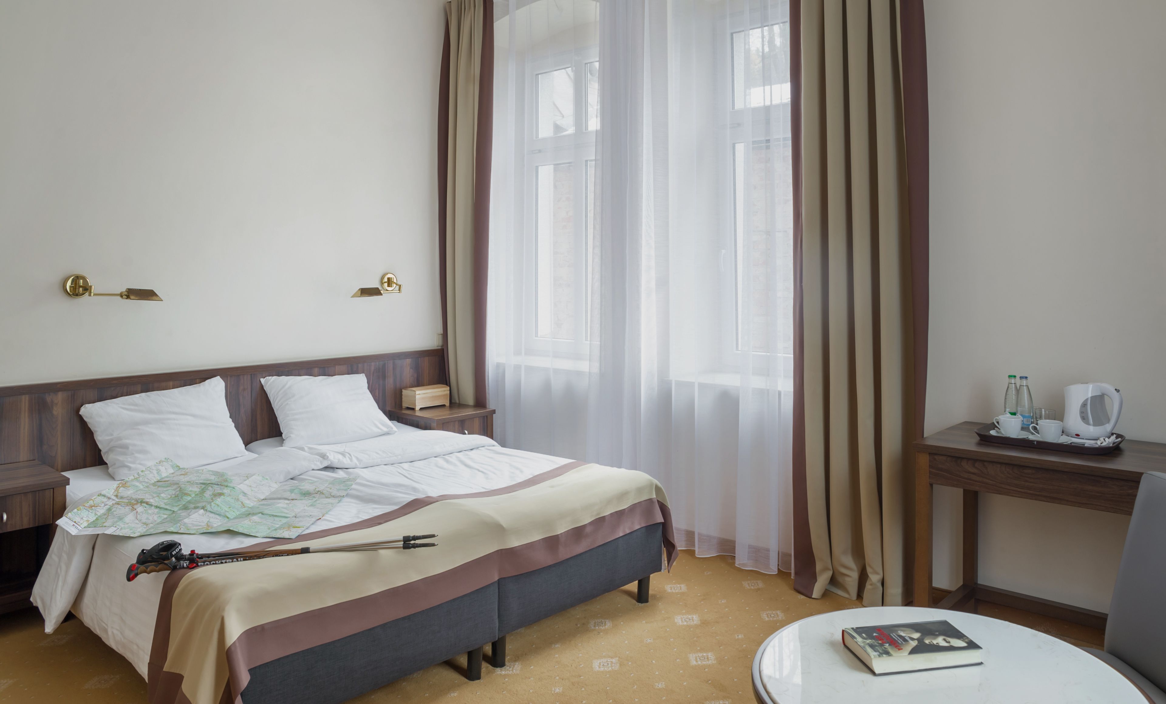 A hotel room with a bed, a bedside table with a kettle, a tray with cups and bottles, and a map on the bed.