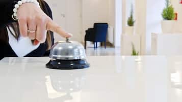 Una mano pulsando un timbre de hotel sobre una mesa blanca en un vestíbulo moderno, bien iluminado, con mostrador de recepción y plantas en macetas.