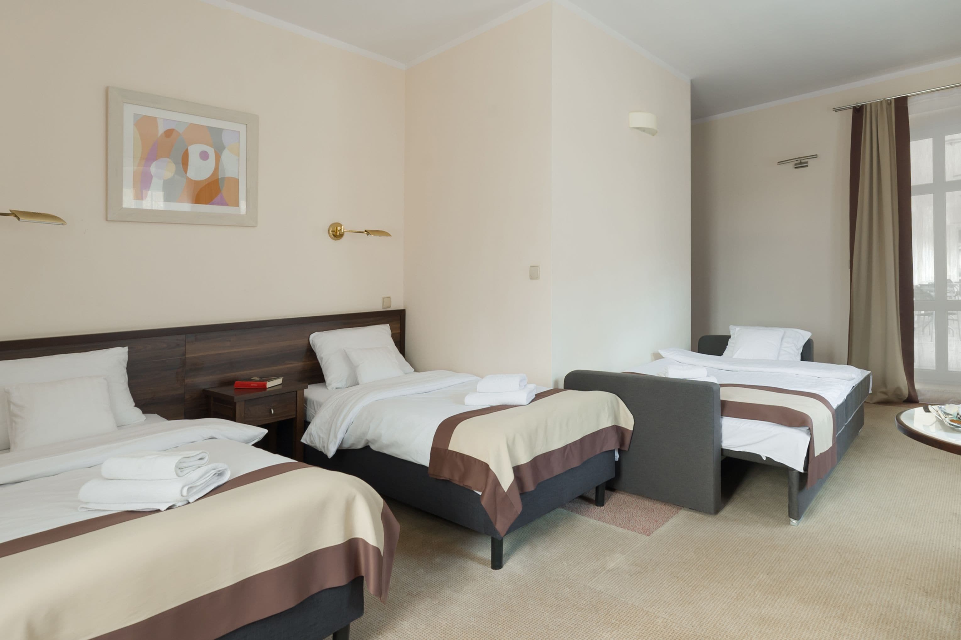 Une chambre d’hôtel avec trois lits, une tête de lit en bois, un tableau abstrait encadré, une table de chevet et une fenêtre avec des rideaux.