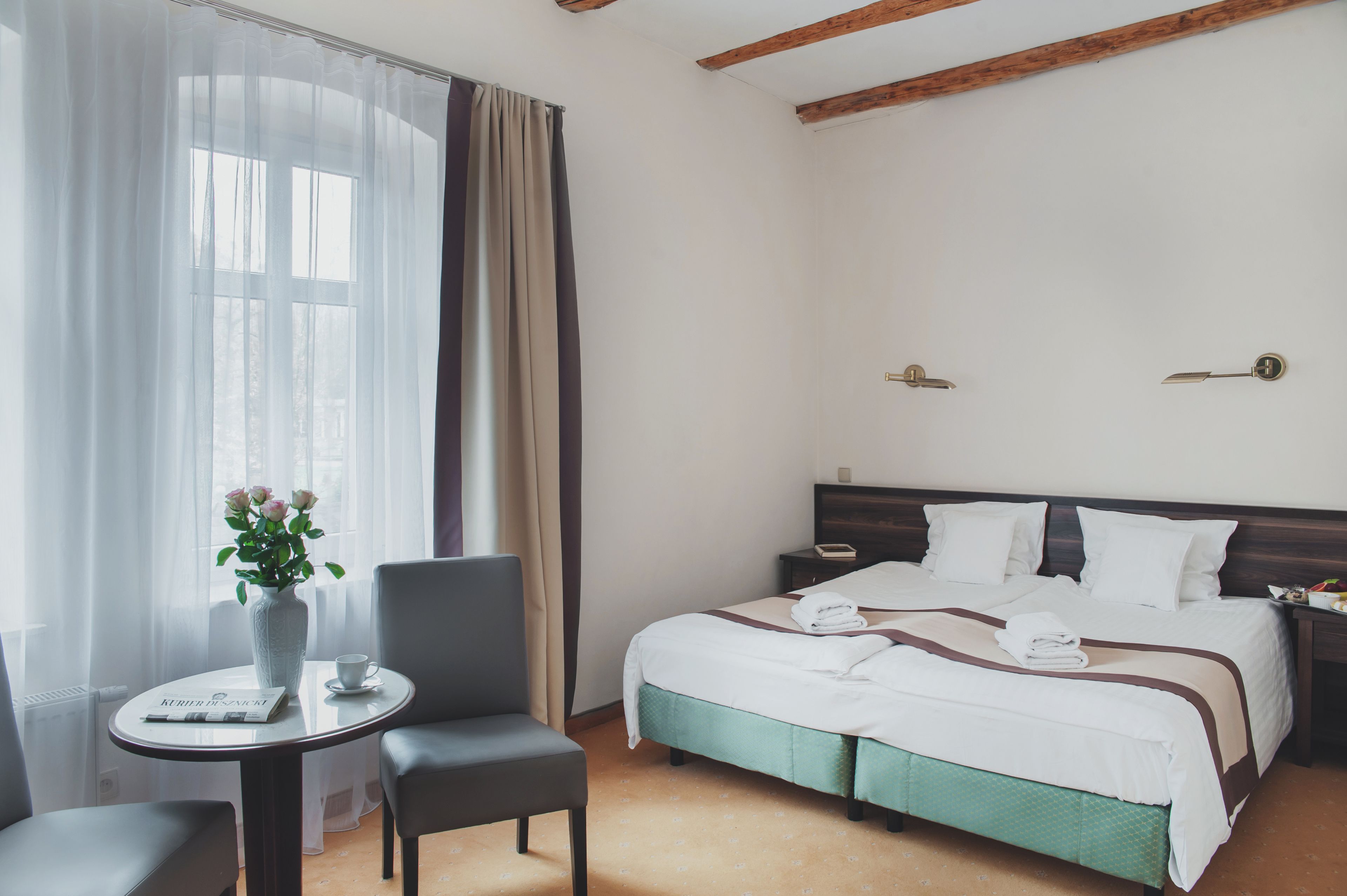 Ett hotellrum med en säng, ett bord med en vas med blommor, en stol och ett fönster med tunna, genomskinliga gardiner.