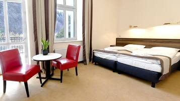 A hotel room with a bed, a red chair, a small table, and a potted plant.