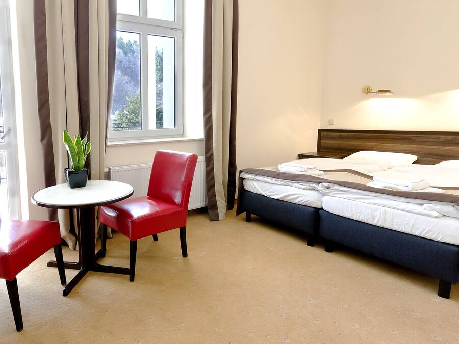 A hotel room with a bed, a red chair, a small table, and a potted plant.