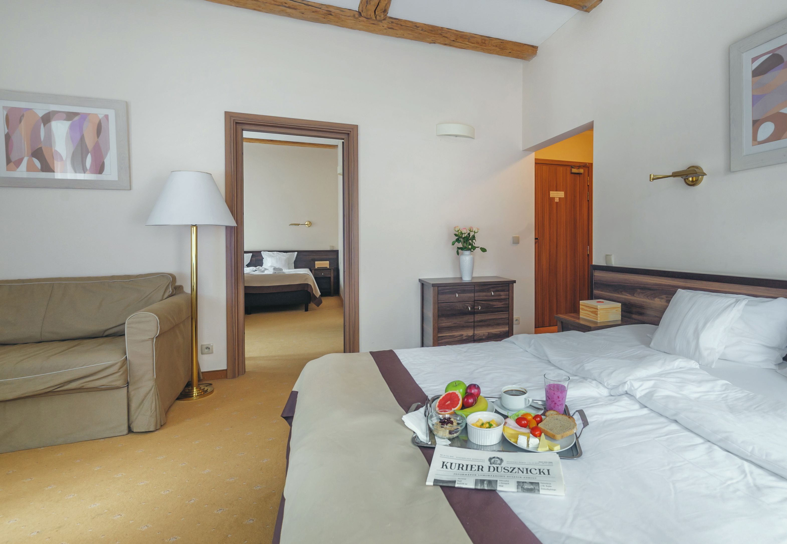 A hotel room with a bed, a bedside table with a lamp, a wooden nightstand, a wooden wardrobe, and a framed picture on the wall.