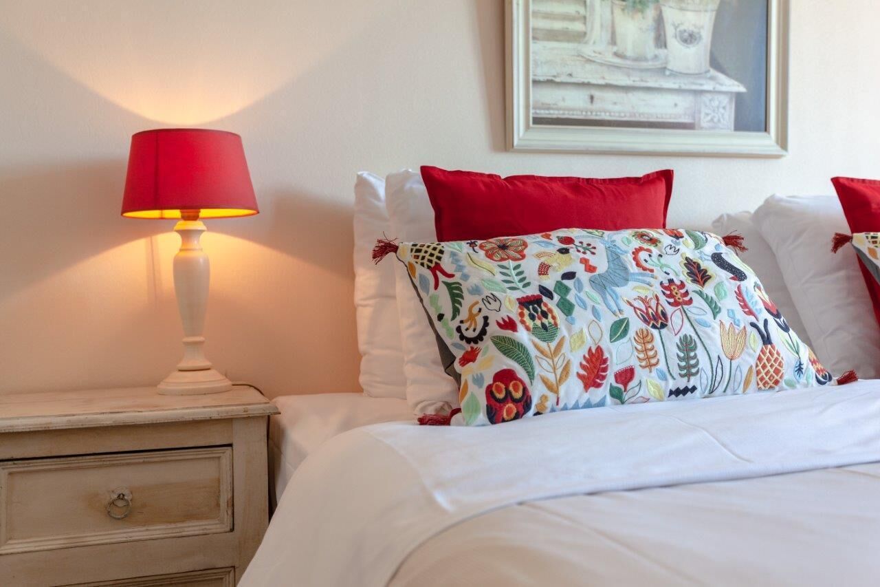 A neatly made bed with white bedding and red pillows, a white bedside lamp, and a colorful embroidered pillow.
