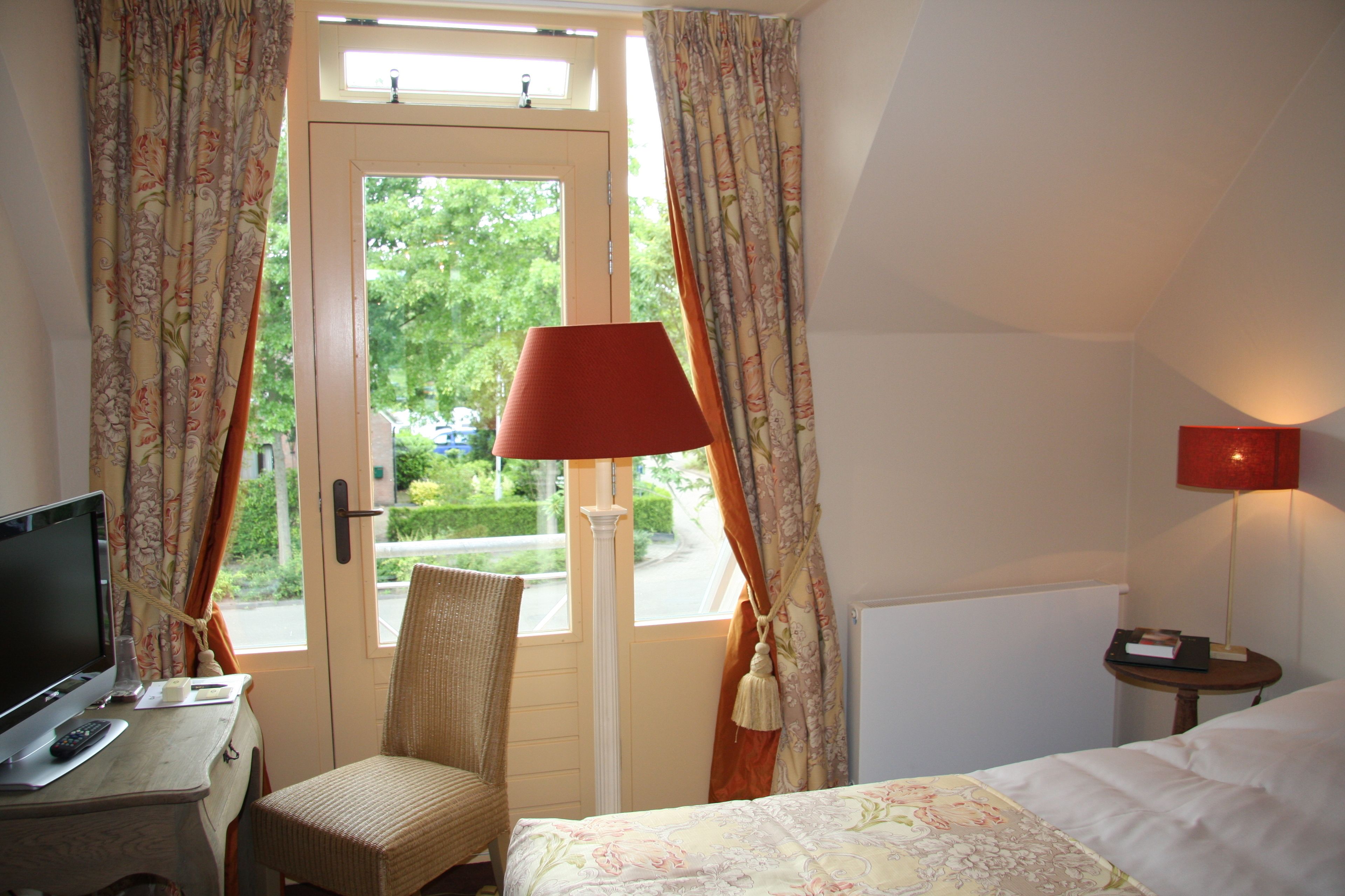 Habitación de hotel con cama, una silla, escritorio con computadora, lámpara y ventana con cortinas.