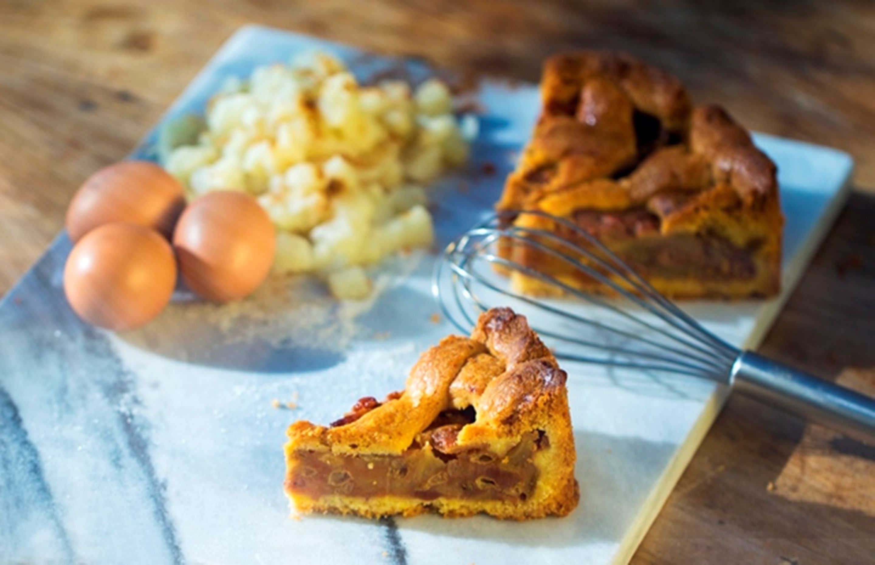 Een stuk appeltaart met een goudbruine korst, een garde en drie eieren op een marmeren oppervlak.