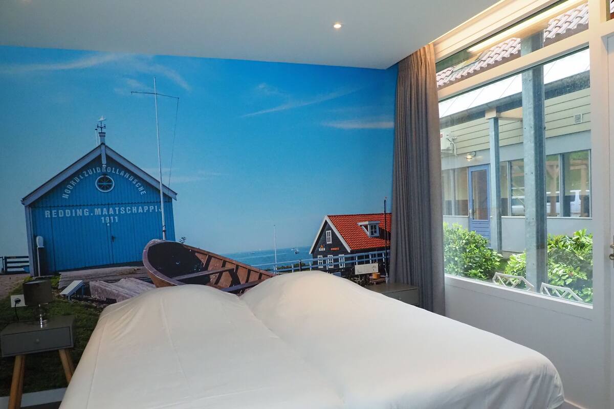 A bedroom with a bed, a window with a view of a blue building and a red-roofed house, and a small table with a lamp.