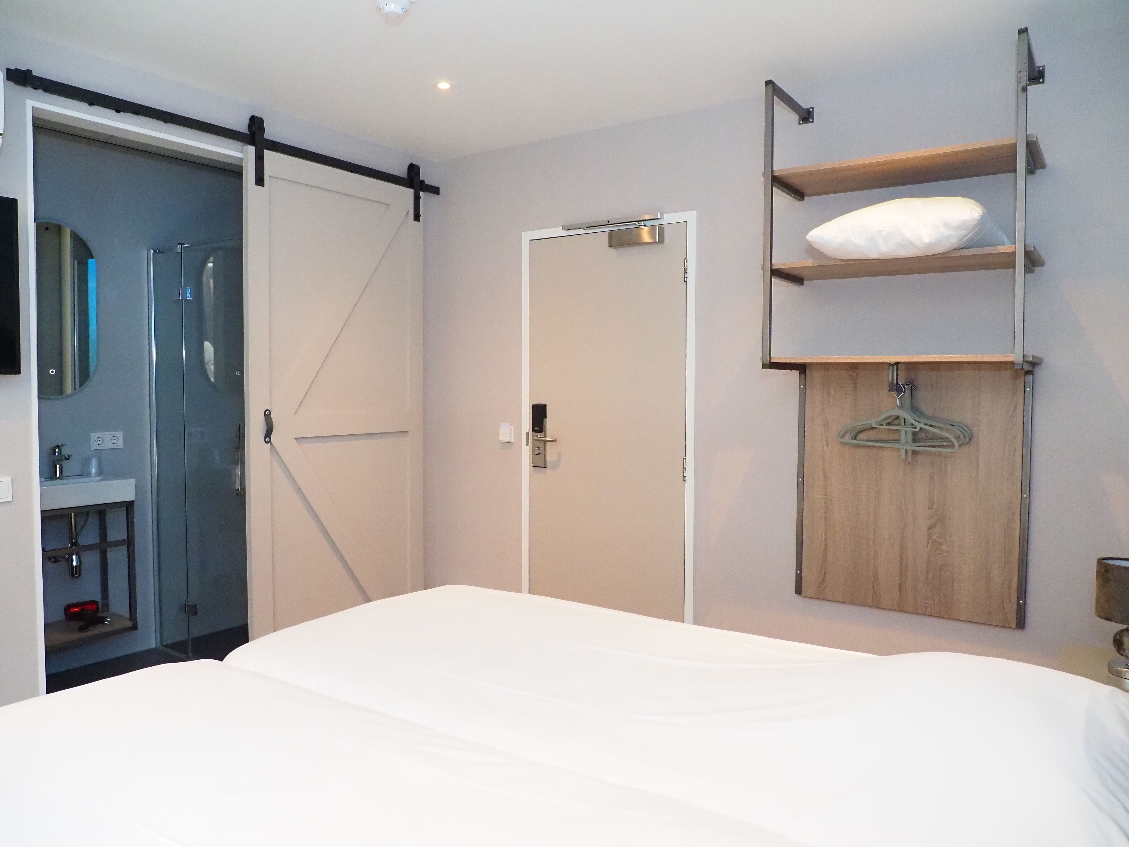 Moderni hotellihuone, jossa on suuri sänky, liukuovi kylpyhuoneeseen, puinen hylly, jossa on tilaa vaatteille, ja pieni yöpöytä.