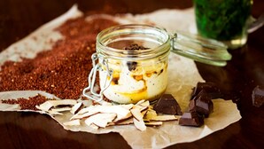 A glass jar with a lid, containing a creamy substance with chocolate pieces, surrounded by scattered chocolate pieces and coconut flakes on a parchment paper. - HarTeluk Joure (Joure)