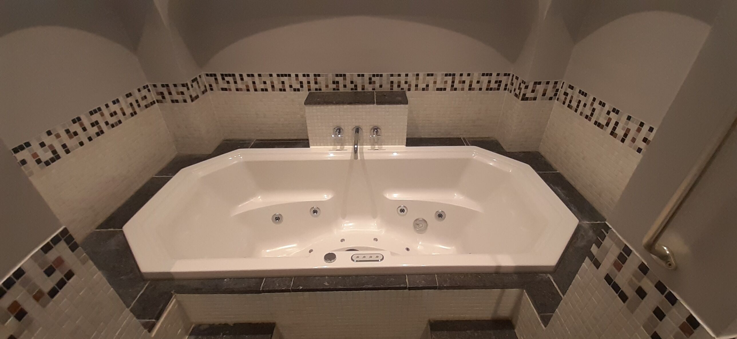 A large, rectangular hot tub with a control panel in the center, surrounded by a tiled wall with a mosaic border.