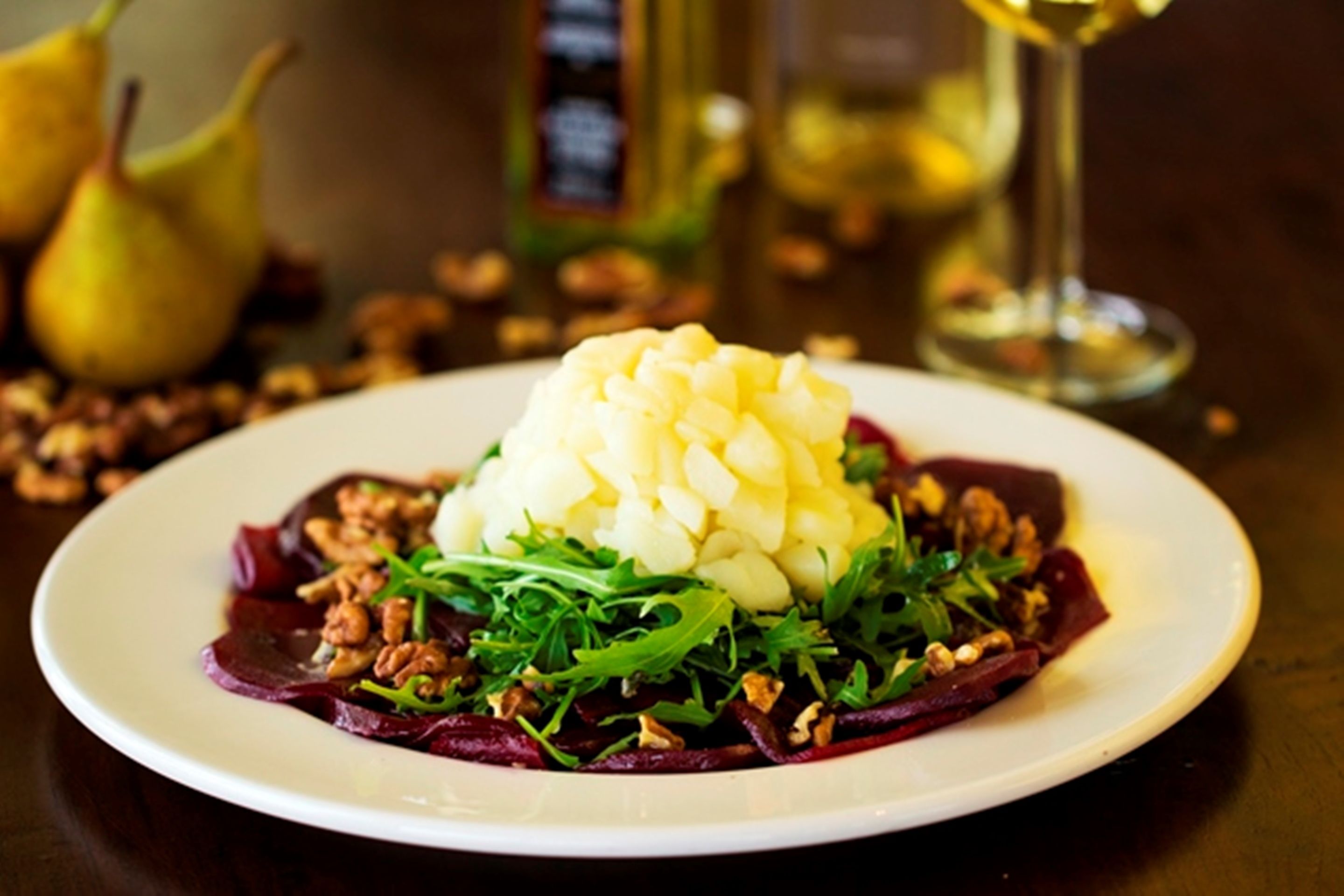 Une assiette remplie de nourriture, avec une portion généreuse de purée de pommes de terre, des légumes verts et des noix, et, au fond, des poires et une bouteille.