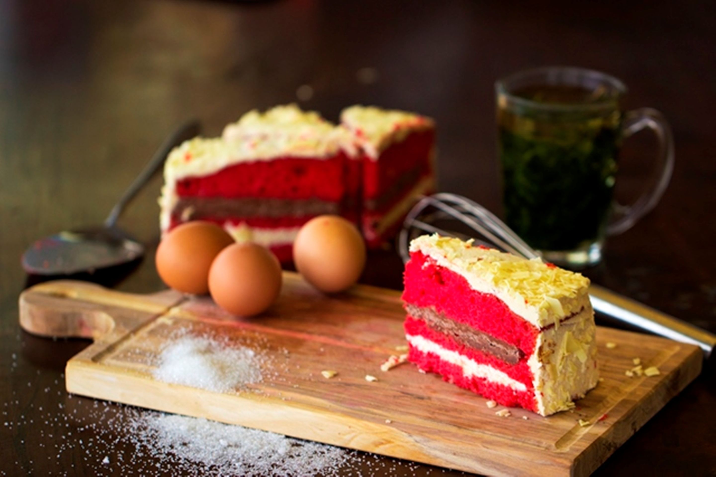 Una rebanada de pastel de terciopelo rojo sobre una tabla de madera con huevos y un recipiente de azúcar cerca.