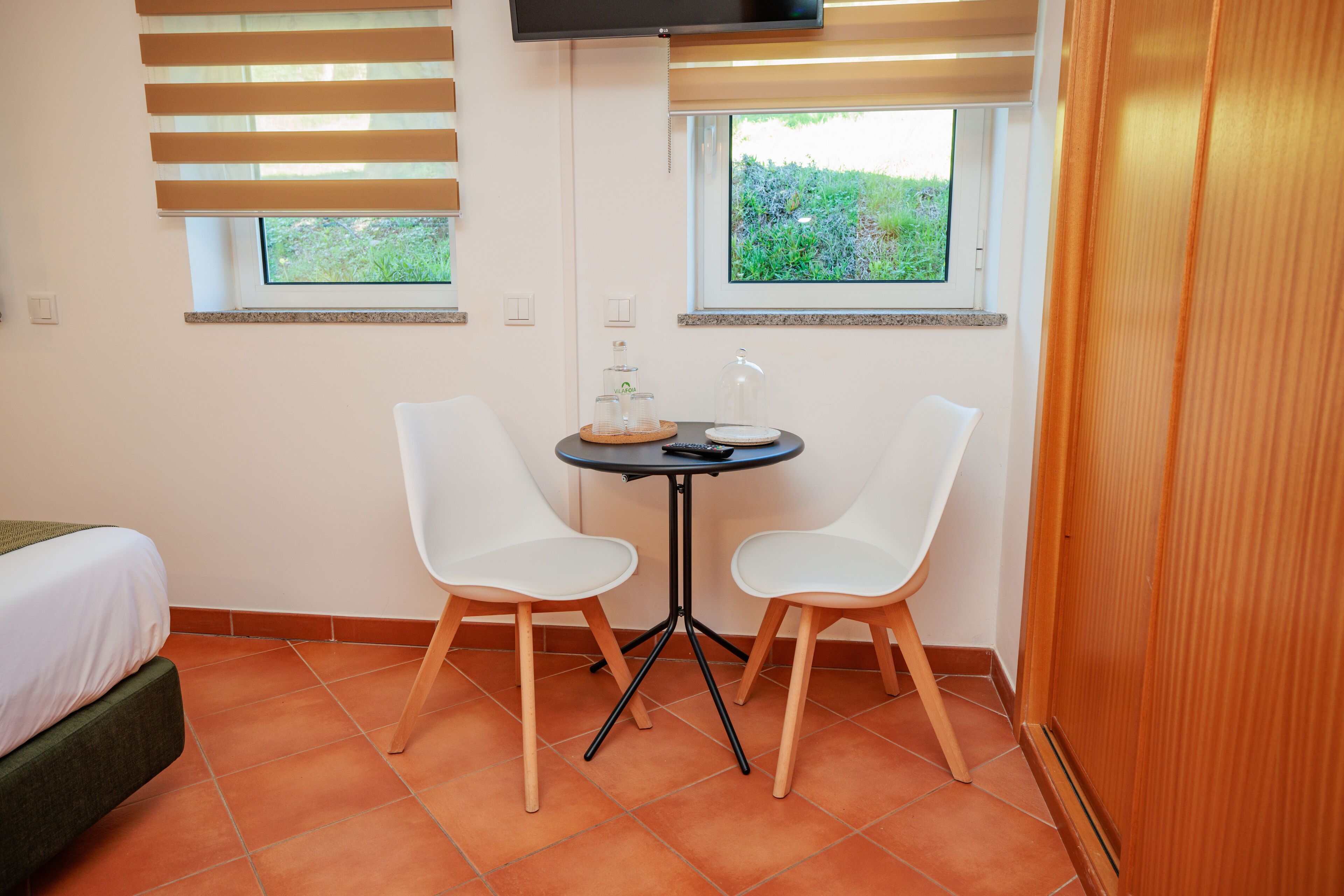 Una habitación pequeña con una cama, dos sillas blancas, una mesita con un recipiente de vidrio y un control remoto, y dos ventanas con persianas.