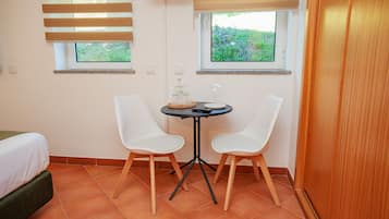 Una habitación pequeña con una cama, dos sillas blancas, una mesita con un recipiente de vidrio y un control remoto, y dos ventanas con persianas.
