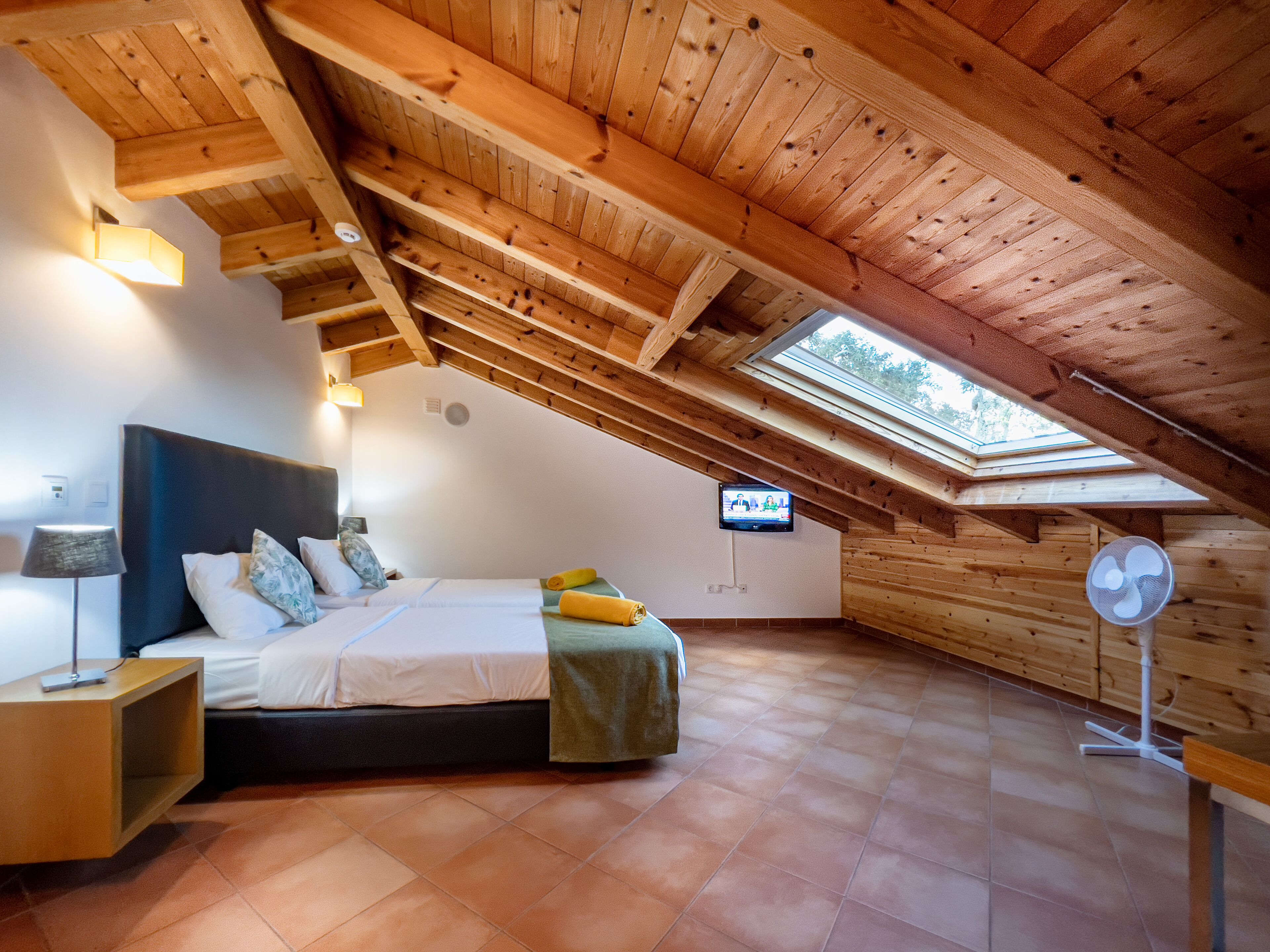 Un dormitorio con techo inclinado, una cama, una mesita de noche, una lámpara, un televisor y un ventilador.