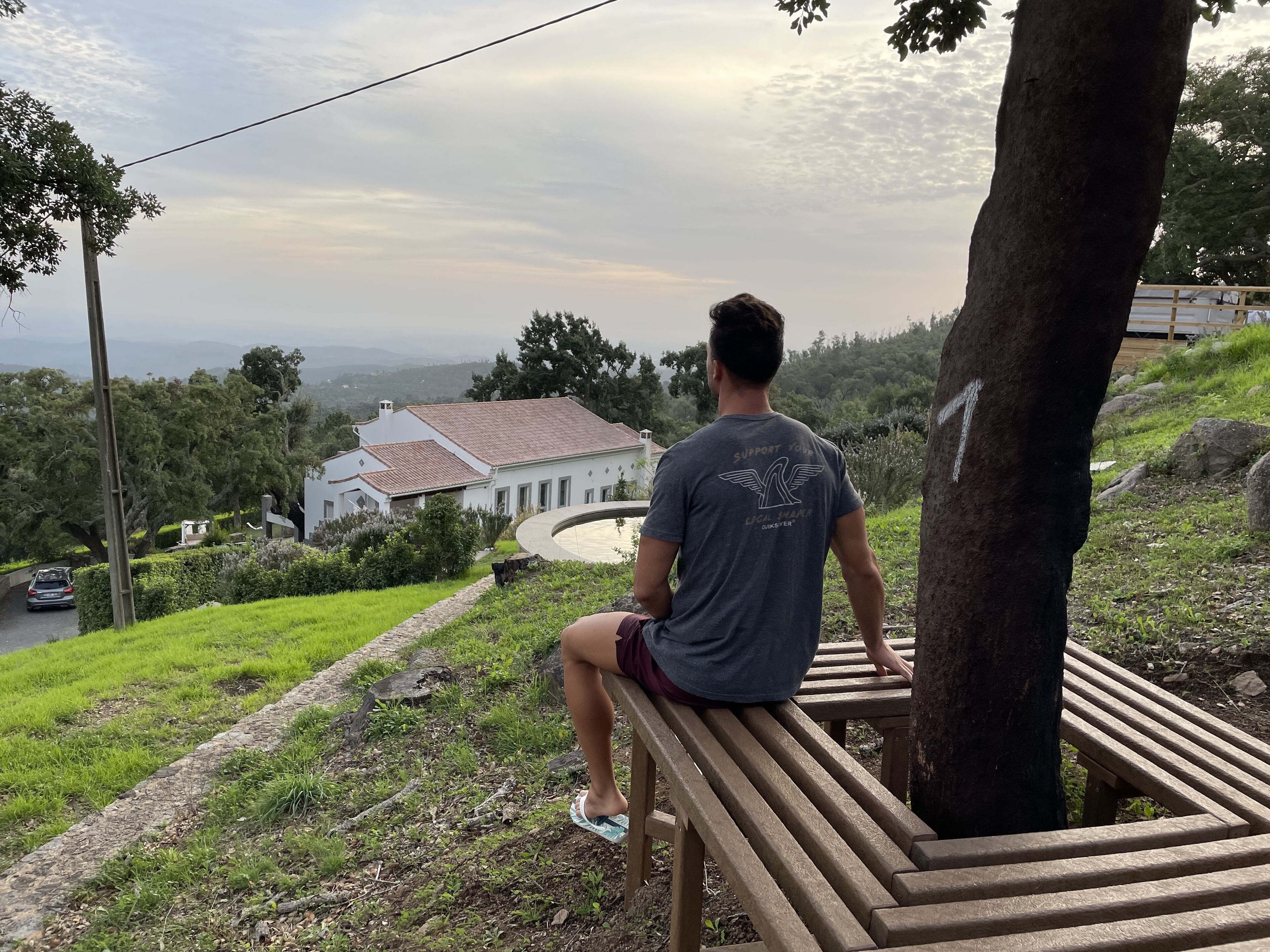A person sitting on a wooden bench overlooking a house with a red roof, surrounded by greenery.