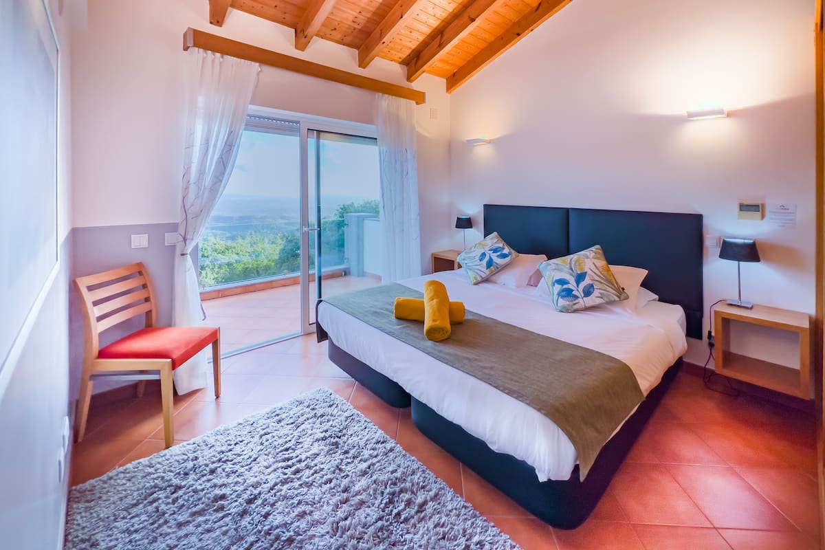 A bedroom with a bed, a chair, a nightstand, a lamp, and a view of the outdoors.