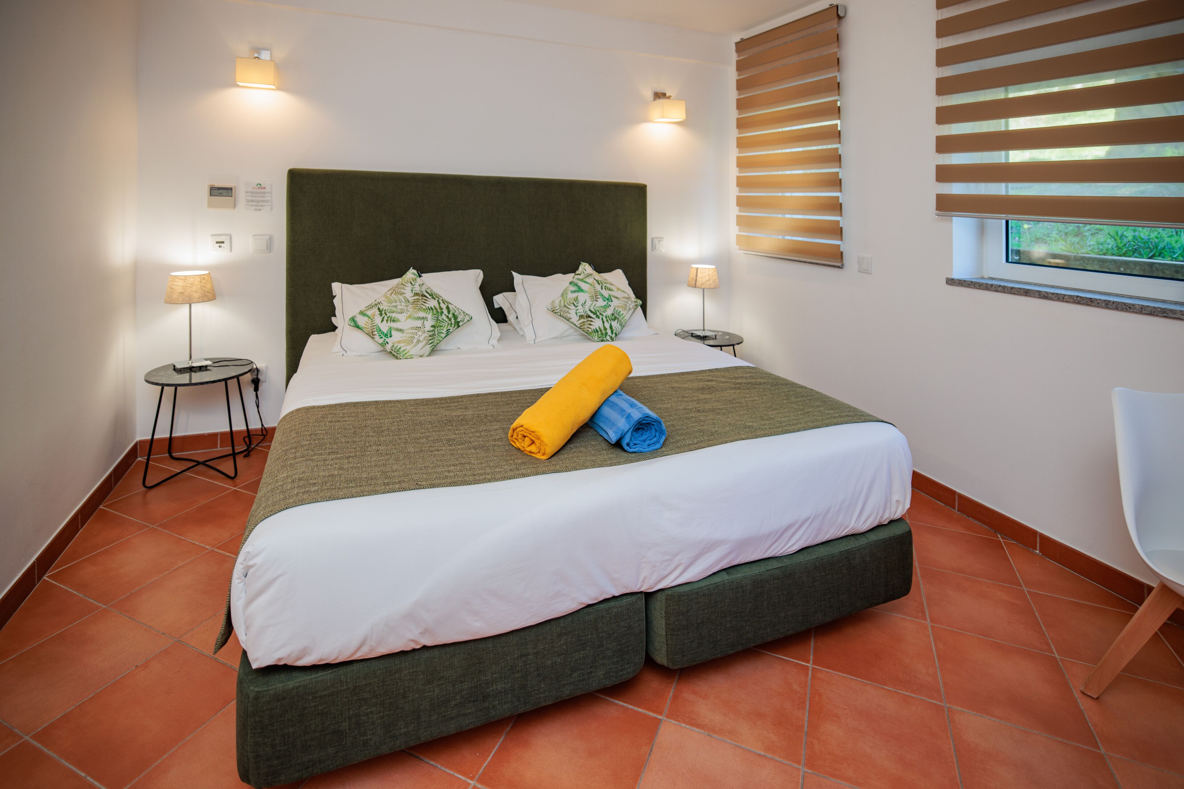 Una cama bien hecha con cabecera verde, ropa de cama blanca y una toalla enrollada. Hay dos lámparas de noche, una ventana con persianas y una silla a la derecha.