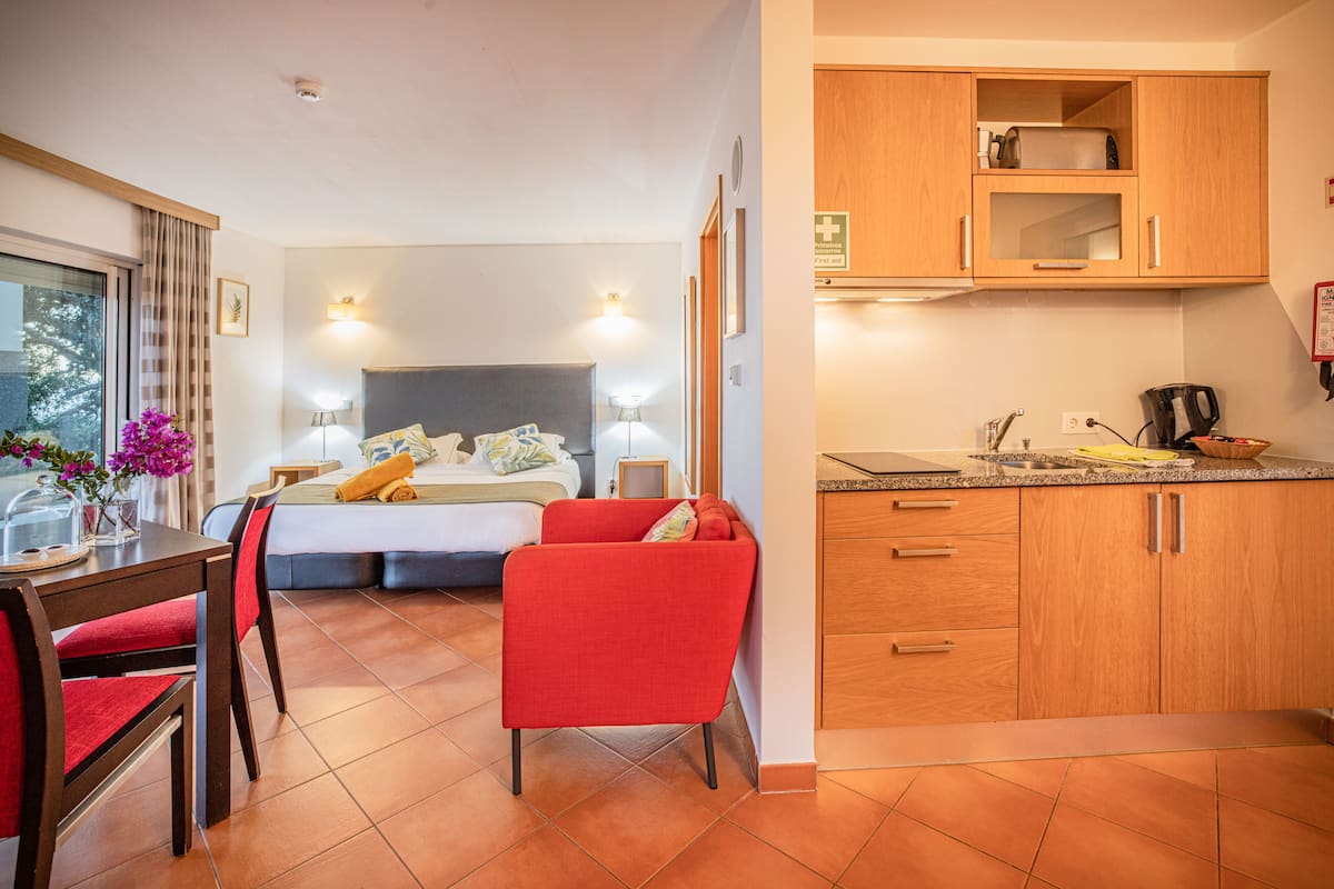 A hotel room with a bed, a red chair, a dining table with chairs, a kitchen area with wooden cabinets, and a window with curtains.